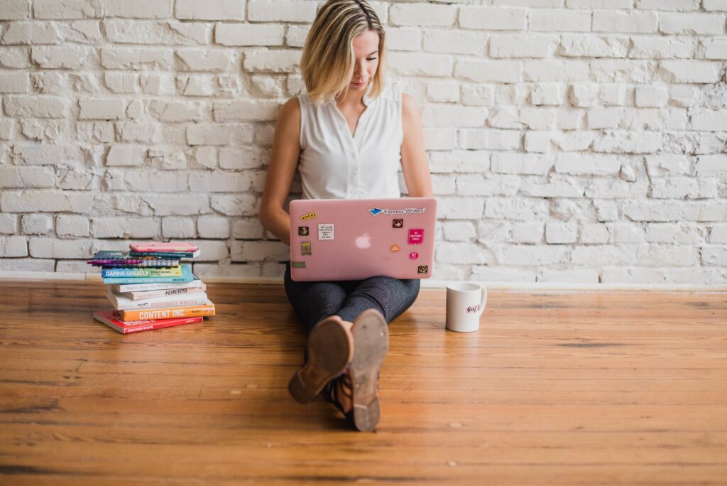 A focused woman seated on the floor, browsing on her sticker-adorned laptop next to a stack of books and a coffee mug, potentially looking to buy cheap papers online